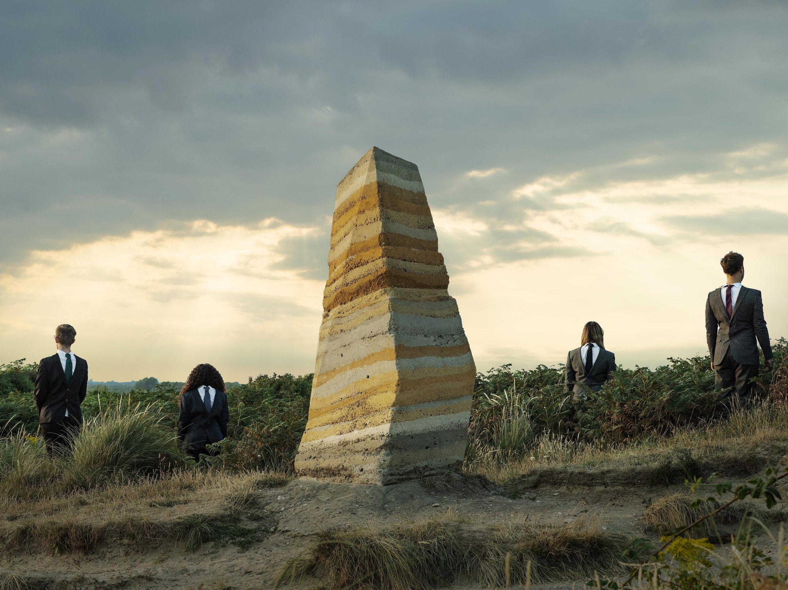 Photograph of four people wearing suits back to front standing around a monolith of rock